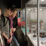 Trzy osoby oglądają wystawę w gablocie, w której znajdują się zbudowane z klocków Lego pierwsze konsole do gier.