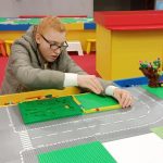 Przy stole z wyłożonymi klockami do zabawy siedzi dziewczynka i tworzy budowlę z klocków Lego.
