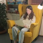Natalia siedzi wygodnie w żółtym fotelu. Czyta tekst. Obok stoi lampa, która ją oświetla.
