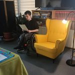 Wojtek czyta swój tekst. Obok stoi żółty fotel i zapalona lampa. Jest skupiony. Siedzi na wózku inwalidzkim.