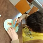 Chłopiec w żółtej bluzce tworzy pracę plastyczna na talerzu. Pochyla się nad stołem, w ręku trzyma mazak, którym dekoruje herbatnik.