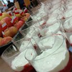 Zbliżenie leżących na stole biało-czerwonych deserów i jabłek z małymi flagami Polski. W tle młodzież uczestnicząca w pikniku.