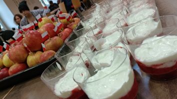 Zbliżenie leżących na stole biało-czerwonych deserów i jabłek z małymi flagami Polski. W tle młodzież uczestnicząca w pikniku.