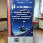 Baner reklamowy Joanny Perlińskiej. Promuje książkę „#RudaRządzi”. Na banerze autorka skacze ze spadochronu. Trzyma ją instruktor.