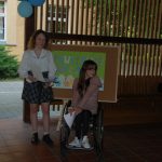 Dwie dziewczęta z Rady Mieszkańców pozują do wspólnego zdjęcia. Jedna stoi i ma w ręku podkładkę ze scenariuszem wystąpienia. Druga siedzi na wózku inwalidzkim. Obie są bardzo zadowolone.