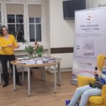 Pani Magda i Pani Aneta stoją i rozmawiają z gościem- Alicją Wiech siedzącą w żółtym fotelu. Na stoliku leżą książki napisane przez Alicję oraz dwa wazony z tulipanami.