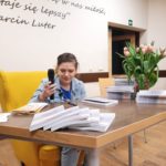 Alicja siedzi przy stole na żółtym fotelu i włącza mikrofon. Na stole leżą książki.