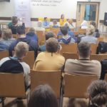 Na krzesłach siedzi publiczność zgromadzona w stołówce na spotkaniu autorskim. W głębi siedzi autorka książki i pani Aneta a obok stoi pani Magda.